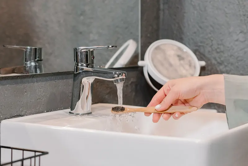 A hand rinsing a bamboo toothbrush under a tap in a modern bathroom sink