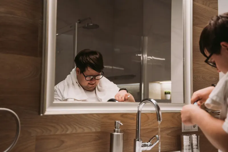 Adult man with glasses reflects in mirror while grooming at bathroom sink with water running