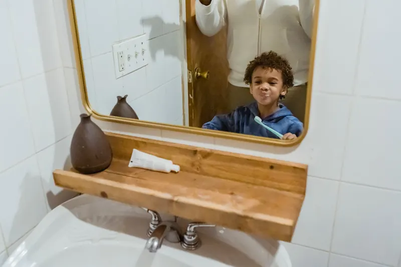 Cheerful African American boy with toothbrush and looking at reflection in mirror while standing in bathroom near sink with anonymous father